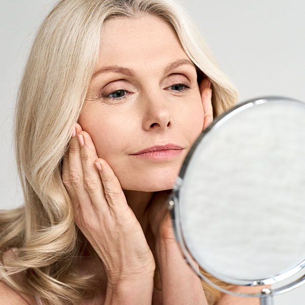Gorgeous happy middle age woman looking at mirror touching her skin enjoying face treatment
